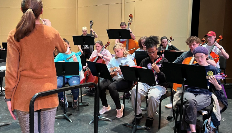 - Instructor conducts a student ensemble during a rehearsal. Students play string instruments and ukuleles while reading from music stands. Double bass players stand in the back row behind seated musicians. The rehearsal takes place on a stage with a simple performance setup.