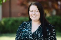 Headshot Victoria Potter - A woman with long dark hair stands outdoors on campus smiling at the camera. She wears a black patterned blouse and a Longwood University name badge. A small nose ring and tattoo near her collarbone are visible. Green shrubs and a brick building appear softly blurred in the background.