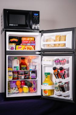 MicroFridge open - A compact refrigerator stands open with a microwave placed on top. The freezer and fridge compartments are filled with frozen meals, yogurt, drinks, and fresh fruit. Beverages and condiments are organized in the refrigerator door shelves. The microwave above features a digital keypad and display.