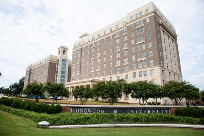 Johns and Moss Halls - Two tall residence hall buildings rise behind a landscaped sign reading “Longwood University.” The buildings feature brick facades, cream stone accents, and many evenly spaced windows. Trees and greenery line the lawn and walkway in front of the complex. A lightly clouded sky stretches above the campus scene.