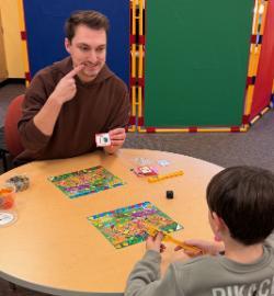 A man in a brown hoodie sits at a round table and plays a colorful board game with a child. He holds up a picture card and points to his mouth as he gives a clue. Game pieces, cards, and a die are spread across the tabletop. Bright blue and green room dividers stand behind them.