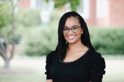 - A woman with braided hair and glasses smiles in an outdoor portrait. She wears a black top and small earrings. Trees and campus buildings appear softly blurred in the background. She faces the camera with a friendly, professional expression.