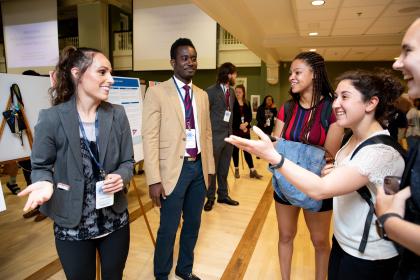 Students participate in Research Day - Students and presenters talk during a research showcase event on campus. Poster presentations are displayed on easels behind them. Participants wear name badges and discuss projects in small groups. The event takes place in a bright indoor hall at Longwood University.