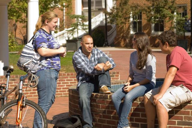 Students sit and talk on brock commons - Four Longwood University students gather and talk around a brick seating area on campus. Two students sit on the low brick wall while two others stand nearby. A bicycle rests beside them as they chat in the sunny outdoor space. Brick buildings and white columns appear in the background.