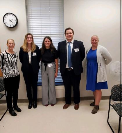 - Five people stand side by side in a classroom, smiling for a photo. Three students wearing business attire and name badges stand between two adults. A wall clock, window with blinds, and classroom chairs appear in the background. The group appears to be attending or presenting at an academic or professional event.
