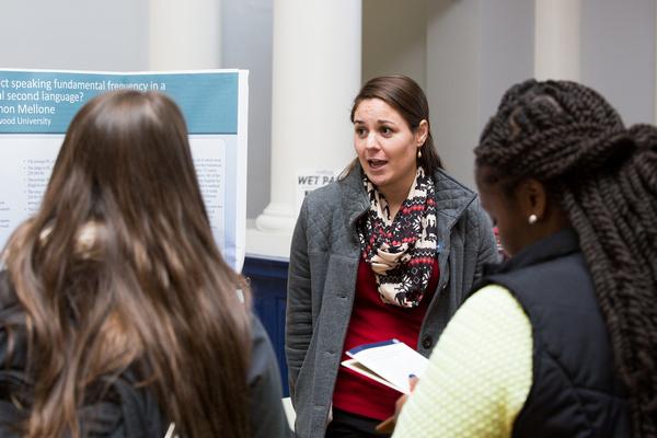 Graduate Poster Session 2014 - A faculty member speaks with two students during a poster session. A research poster is displayed on a board beside them in an academic setting. The students listen while one holds a notebook and pen.