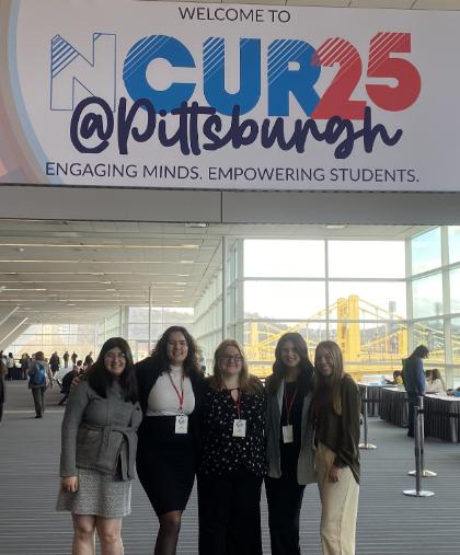 - Five students stand together beneath a sign reading “Welcome to NCUR25 @ Pittsburgh.” They wear conference badges and pose inside a large convention center. A yellow bridge and city view are visible through the windows behind them. Students attend and represent their university at a national research conference.