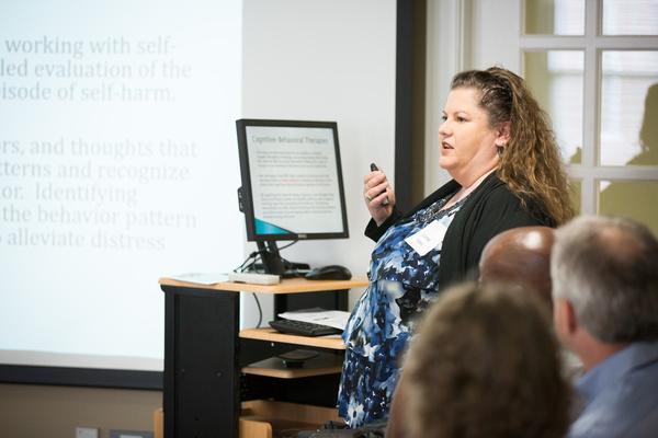 Grad studies Presentation - A presenter speaks to an audience while holding a remote during a lecture. A slide behind her includes text about self-harm and evaluation strategies.