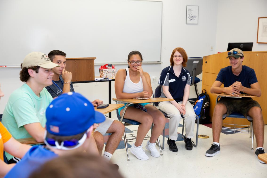 Students and coach during their first coaching group meeting - Students sit in a circle of desks during a classroom discussion. Several students smile and listen while talking with one another. A student leader sits among them facilitating the conversation. Group discussion takes place in a campus classroom setting.