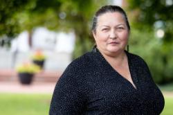 Beth Mottley - Woman with gray-streaked hair stands outdoors on Longwood’s campus. She wears a black blouse with small white dots and dark earrings. Trees and greenery blur softly in the background. Portrait-style photo taken in natural daylight.