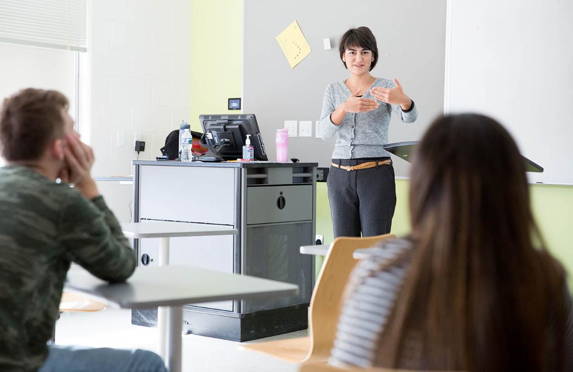 Longwood professor teaching students in class - An instructor stands at the front of a classroom and gestures while speaking. Two students sit in the foreground, listening from their desks. A lectern and instructor station with a computer sit beside a whiteboard. Daylight from a window brightens the room.