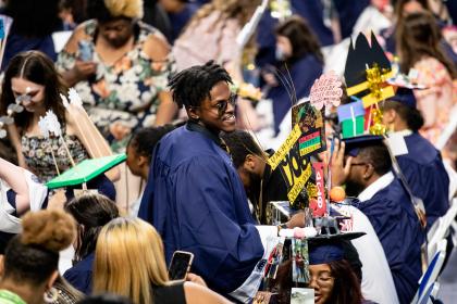 A student without a cap on is looking around and smiling at everyone else being capped in the Joan Perry Brock Center at the 2023 Convocation - Graduate in cap and gown smiles while standing among seated classmates. Decorated mortarboards with signs, photos, and colorful designs fill the crowd. Families and guests watch and take pictures from nearby seats. The ceremony setting is busy with graduat