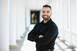 Waleed Ahmed - A man smiles in a professional portrait in a bright hallway. He wears a black button-down shirt and stands with his arms crossed. He faces the camera with short curly hair and a trimmed beard. White columns and windows create a clean, softly blurred background.