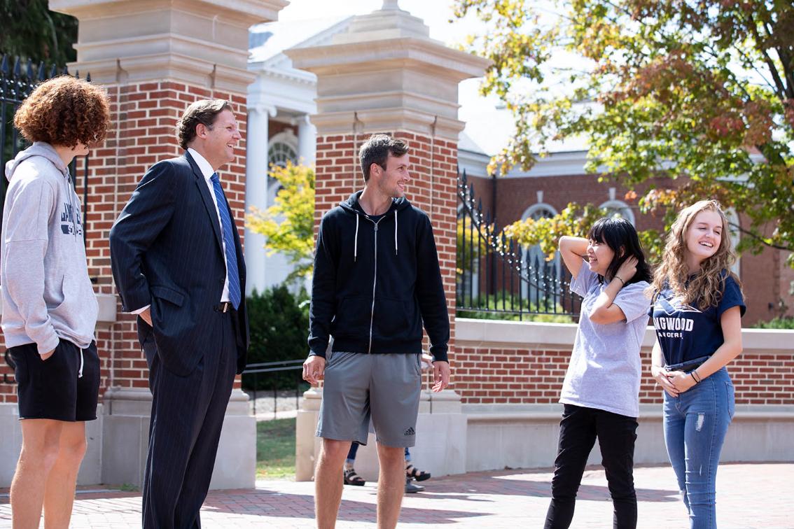 President Reveley talking with students on campus - A group of students talk and laugh with a staff member outdoors on campus. They stand on a brick walkway beside tall brick pillars and an iron gate. Fall trees with green and orange leaves frame the scene in bright sunlight. One student wears a Longwood Lancers T-shirt as the group chats.