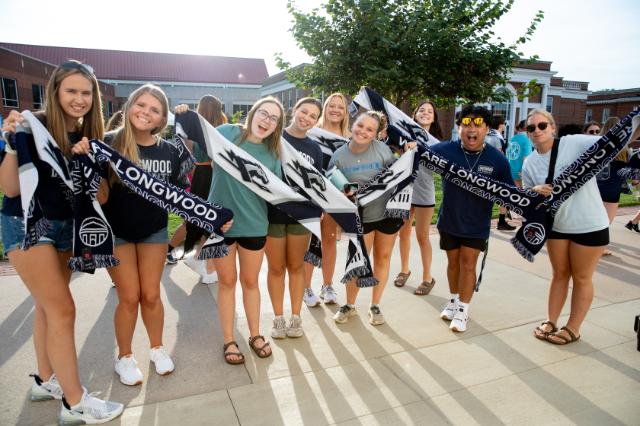 Students with their scarves - Group of Longwood students pose outdoors on campus during a spirit event. They hold blue-and-white “We Are Longwood” scarves with the Lancer logo. Students smile and cheer in the afternoon sun near campus buildings and a tree. Other attendees and walkways appear in the background.