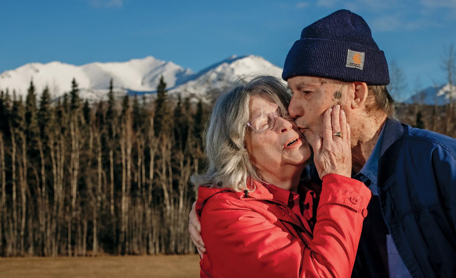 - An older couple stands outdoors with snow-covered mountains in the distance. The woman in a red jacket gently touches the man’s face. He wears a blue jacket and knit cap as they share a kiss. Evergreen trees and a clear sky frame the scenic backdrop.