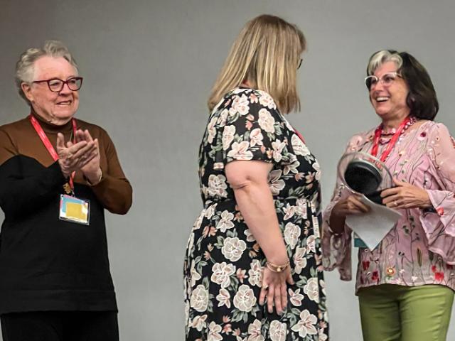 Dr. Lily Anne Goetz receives the FLAVA Dr. Helen Warriner-Burke Distinguished Service Award from FLAVA President Heidi Trude as Warriner-Burke looks on. The award is named for Warriner-Burke '56. - Three women stand together during a recognition event. One woman smiles while holding an award and papers. Another woman faces her as a third applauds from the side. All wear name badges, suggesting a c