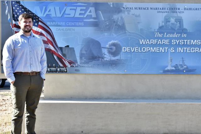 Joe Gills '15 - A man stands beside a large sign for the Naval Surface Warfare Center in Dahlgren. An American flag and images of naval ships appear on the sign behind him. Text on the display references warfare systems development and integration. He wears a button-down shirt and slacks outdoors in front of the facility.
