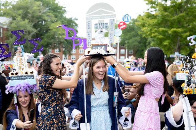 Kara Parr '23 is capped by friends - Two friends adjust a graduate’s decorated mortarboard during commencement. The cap features large purple letters and a tall elementary education display. The graduate smiles while standing among a crowd of classmates. Brick residence halls and trees fill the background.
