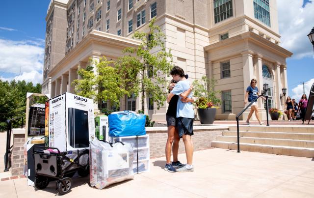 A couple hugging outside one of the high rise residence halls - Two people embrace outside a residence hall during move-in day. Boxes, storage bins, a mini fridge, and a wagon of belongings sit nearby. The tall brick and cream-colored building rises behind them. Other families walk up the steps under a bright blue sky.