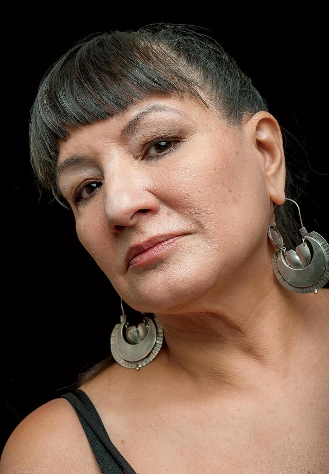 Sandra Cisneros (Photo credit: Keith Dannemiller) - Close-up portrait of a woman against a black background. She wears large silver circular earrings and a black top. Her dark hair is pulled back with straight bangs framing her face. She looks slightly to the side with a calm, thoughtful expression.