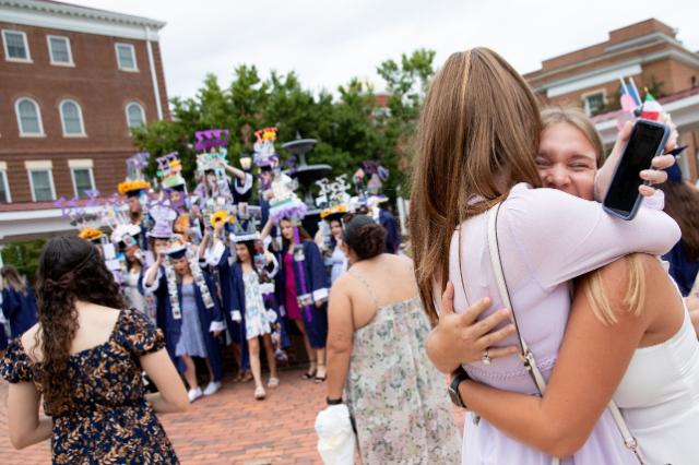 Two students hug after convocation beside the fountain where a large group is posing for a photograph - Two women embrace during a graduation celebration on campus. One smiles with eyes closed while holding a phone in her hand. Graduates in caps and gowns with decorated mortarboards gather behind them. Brick buildings and a fountain frame the festive scene.