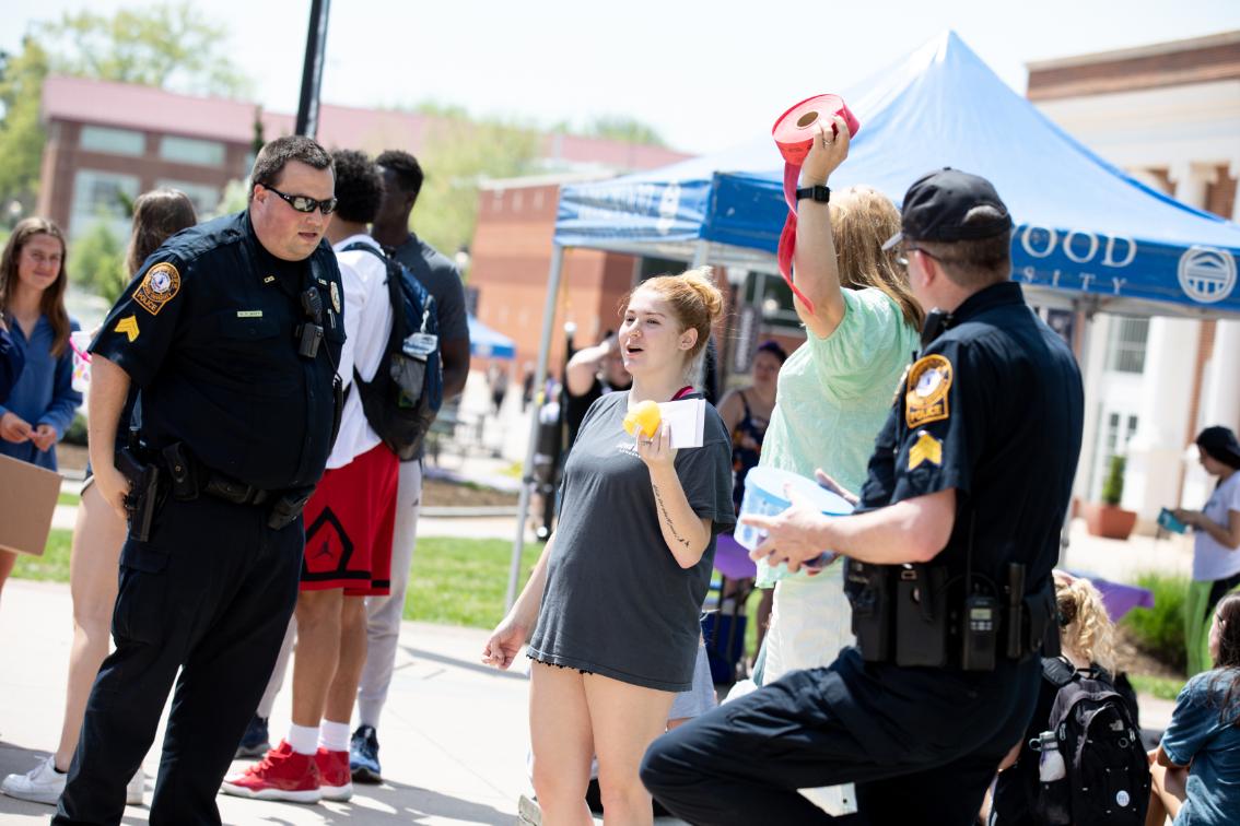 Officers at the LUPD annual egg hunt - Campus police officers talk with students at an outdoor event. One student holds a yellow plastic egg while another raises a roll of red tickets. A blue Longwood University tent stands behind them. Other students gather on the lawn on a sunny day.