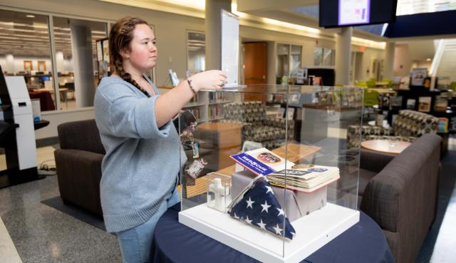 A flag that flew at Ground Zero was displayed at Longwood this week, brought to campus by our associate vice president of information technology services, an Air Force veteran. - A woman places a clear cover over a display case in a campus library. Inside the case are a folded American flag, a small white cross, and a book titled “Rescue.” The display sits on a table surrounded by lounge seating a