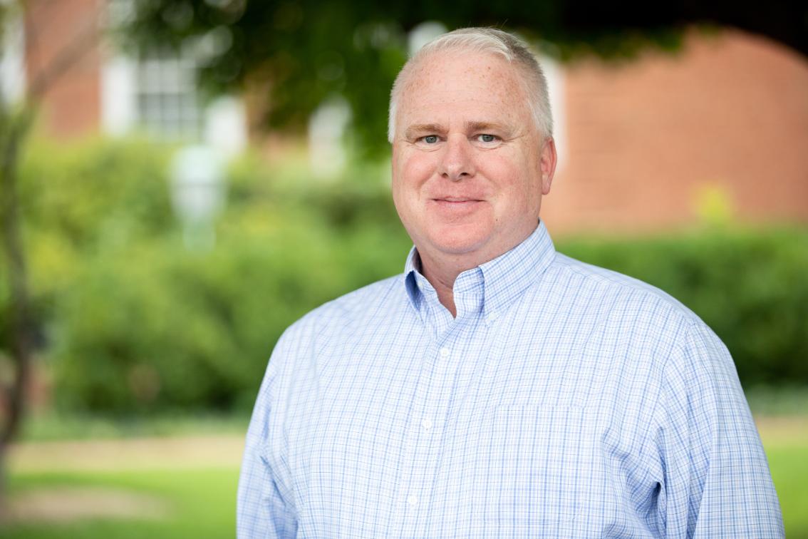 Jason Ferguson (M.S. ‘12) - Man with short gray hair smiling outdoors on campus. He wears a light blue checkered button-down shirt. Green shrubs and trees fill the softly blurred background. A brick building is visible behind him.