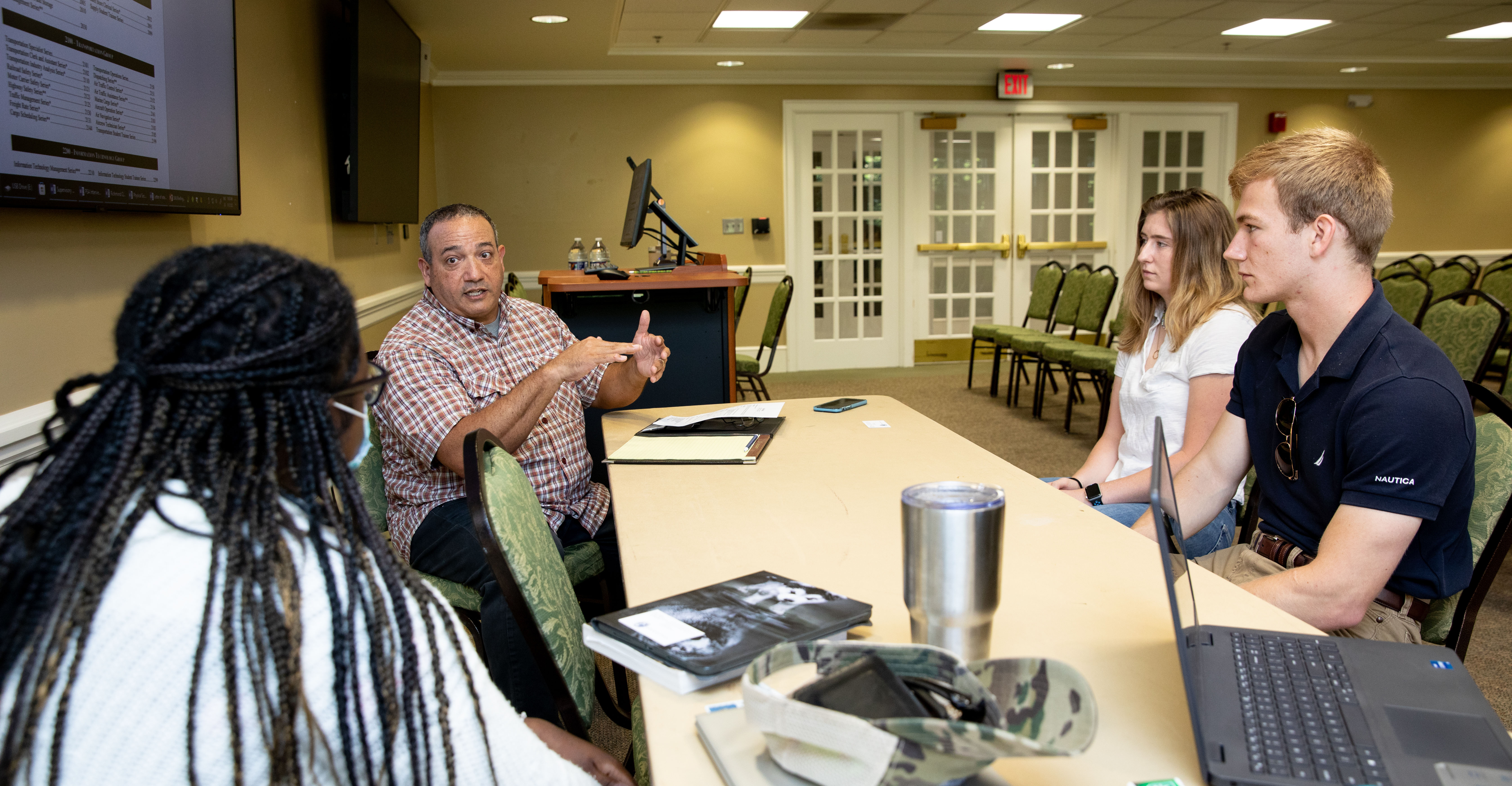 Jamie Finney ’98 works with current students - A professor speaks to three students seated around a conference table. He gestures while discussing material displayed on a screen in the room. Students listen attentively with a laptop and notebooks open. The meeting takes place in a campus classroom or seminar space.