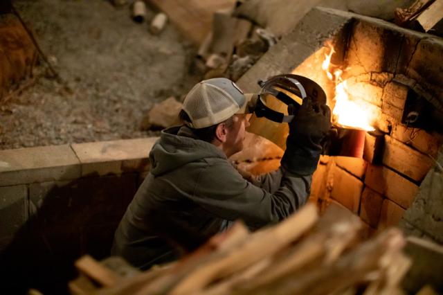 Person tending to the kiln - Man wearing gloves and a cap peers into a brick kiln filled with flames. He lifts a protective face shield while checking on a ceramic piece inside. Bright fire glows against the stacked bricks of the outdoor kiln. Wood logs rest nearby as he carefully monitors the firing process.
