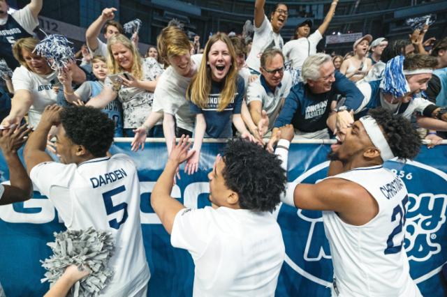 Basketball players greet fans after a game - Longwood men’s basketball players celebrate with fans after a game. Players in white jerseys reach up to high-five cheering supporters over a railing. Fans wearing Longwood shirts lean forward with raised arms and pom-poms. The arena crowd appears energized and joyful.