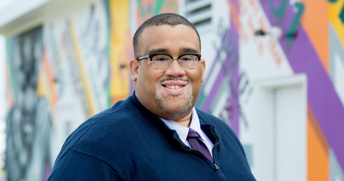Cameron Patterson ’10, Interim Vice President for Student Affairs - Portrait of a man wearing glasses, a navy sweater, and a tie. He stands outdoors in front of a colorful mural. The background features abstract shapes and bright painted panels. He smiles toward the camera.