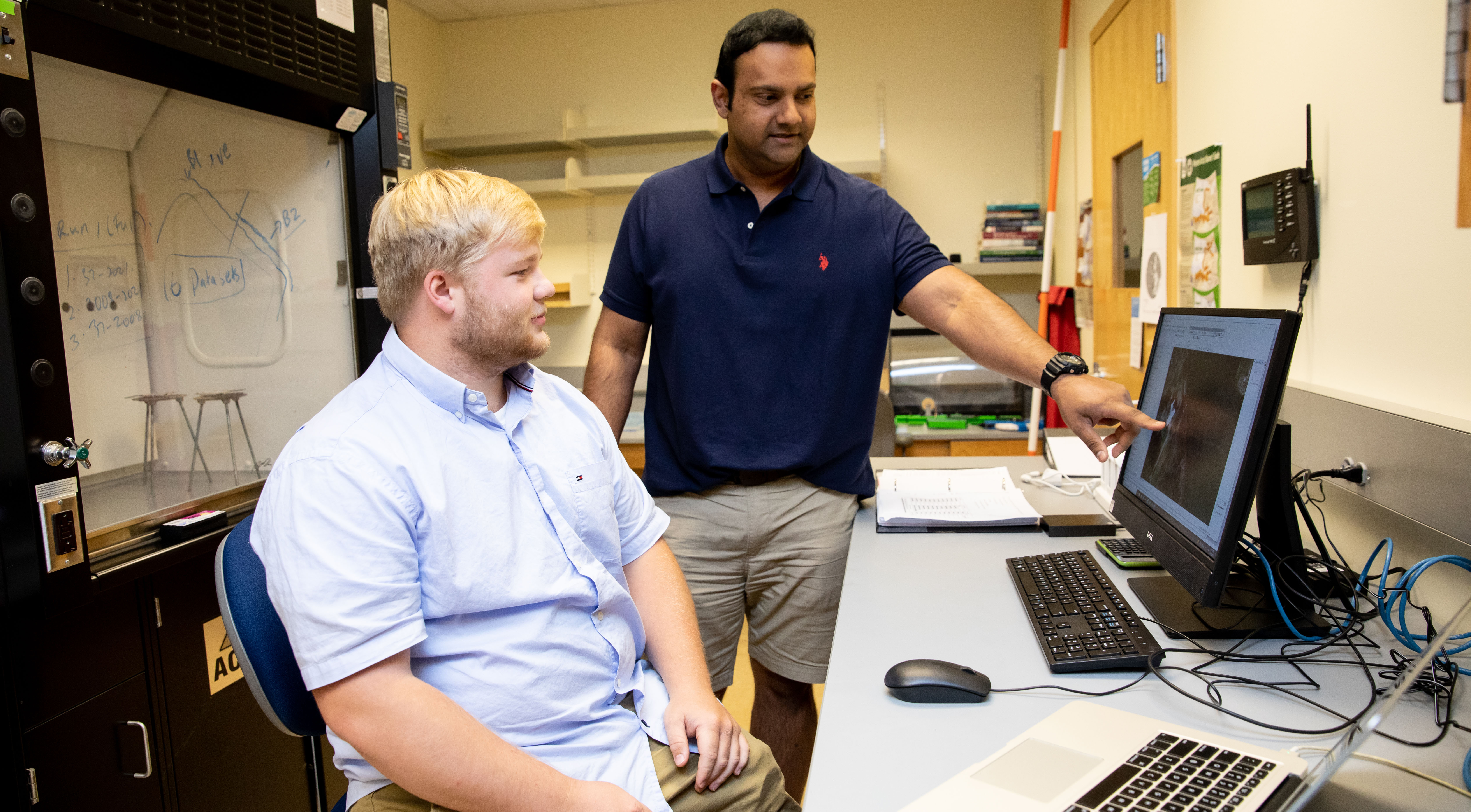 Matthew Wright ‘23 working with Dr. Ravi Darwin Sankar - Professor points to data on a desktop monitor while a student looks on. They stand and sit at a lab workstation with keyboard and mouse. A fume hood and whiteboard are visible in the background. The scene shows one-on-one instruction in a science lab setting.