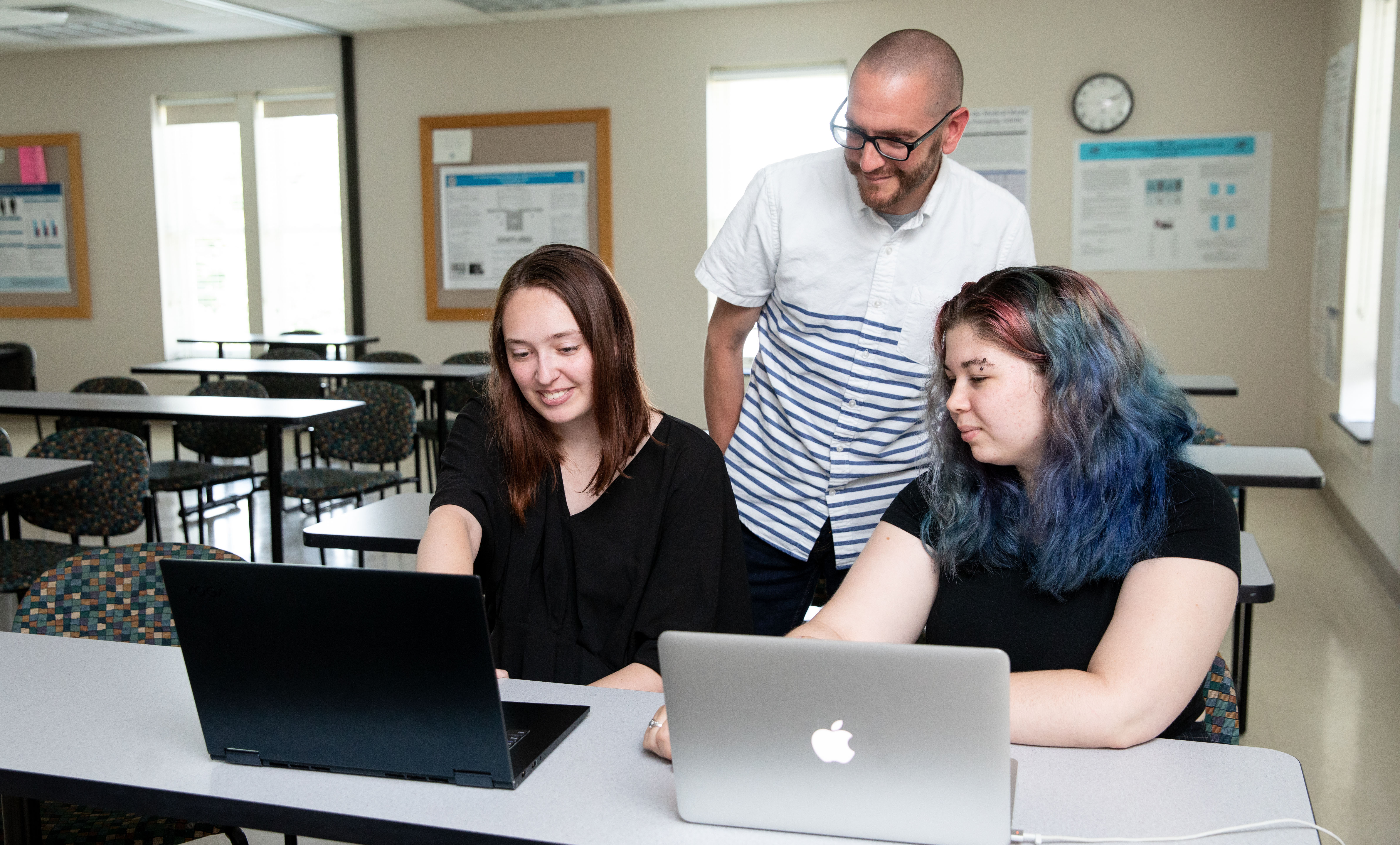 Dr. Timothy Ritzert with Kylie Gannon ‘23 and Leah Richardson ‘23 - Two students sit at desks working on laptops in a classroom. An instructor stands behind them offering guidance. The students look at their screens while discussing their work. Tables, chairs, and research posters line the room.