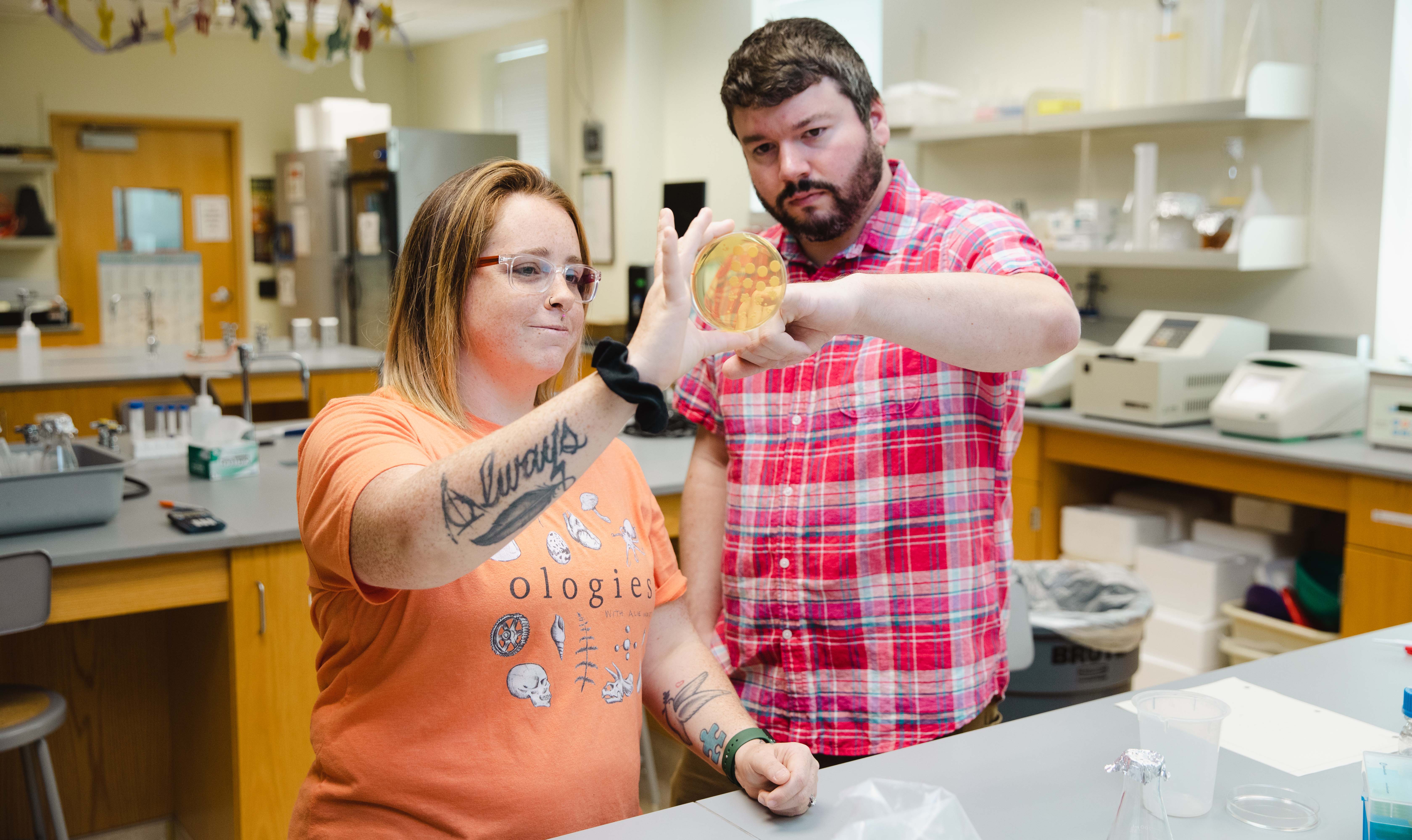 Jammie McMurtry ’24 working in the lab with Dr. Jonathan White - Two researchers examine a petri dish in a science laboratory. One holds the dish up to the light while the other looks closely at the sample. Lab equipment and instruments line the counters behind them. The scene highlights hands-on scientific research and collaboration.