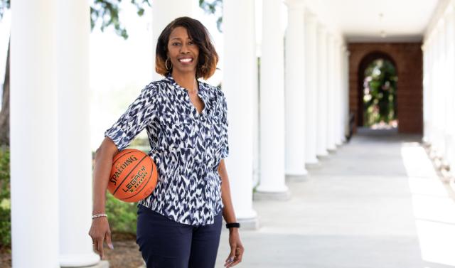 Erika Lang-Montgomery - Woman standing beneath white campus columns holding a basketball at her side. She wears a patterned blouse and navy pants, smiling at the camera. The orange Spalding basketball rests against her hip. A brick archway and greenery appear in the background.