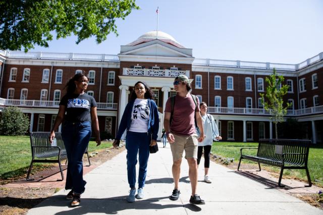 Students walking to class from the Rotunda - Four students walk along a campus sidewalk on a sunny day. They talk and smile in front of a red brick building with white columns and a dome. Benches and green lawns line the pathway. Trees frame the scene under a clear blue sky.