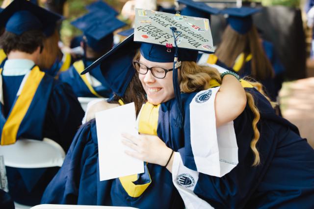 Students hug during the commencement ceremony - Two graduates in blue caps and gowns embrace during a commencement ceremony. One graduate smiles while holding a program in her hand. Her decorated mortarboard features handwritten messages and small drawings. Other graduates in regalia sit in rows in the background.