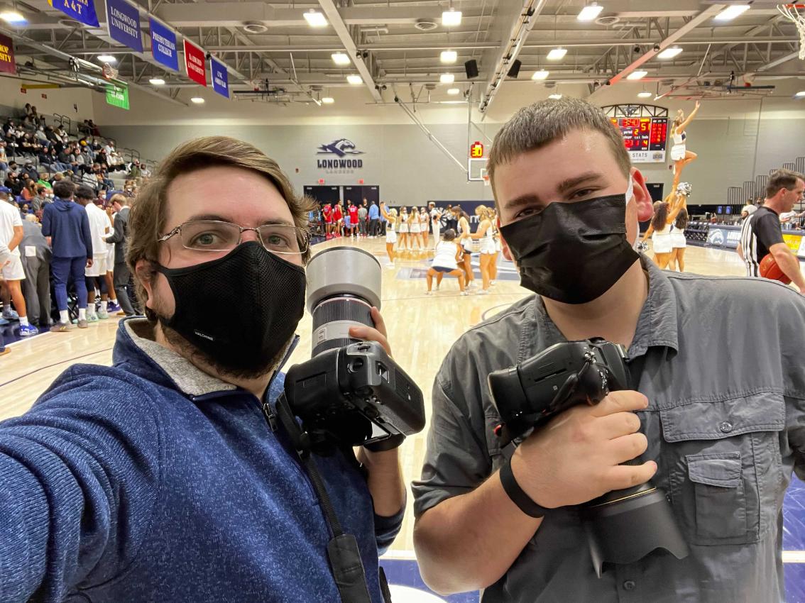 Mike Kropf ’14 and Connor Thompson ’23 - Two photographers wearing face masks pose courtside at a Longwood basketball game. Each holds a camera with a large telephoto lens. Cheerleaders perform on the court behind them. Fans fill the stands inside the arena.