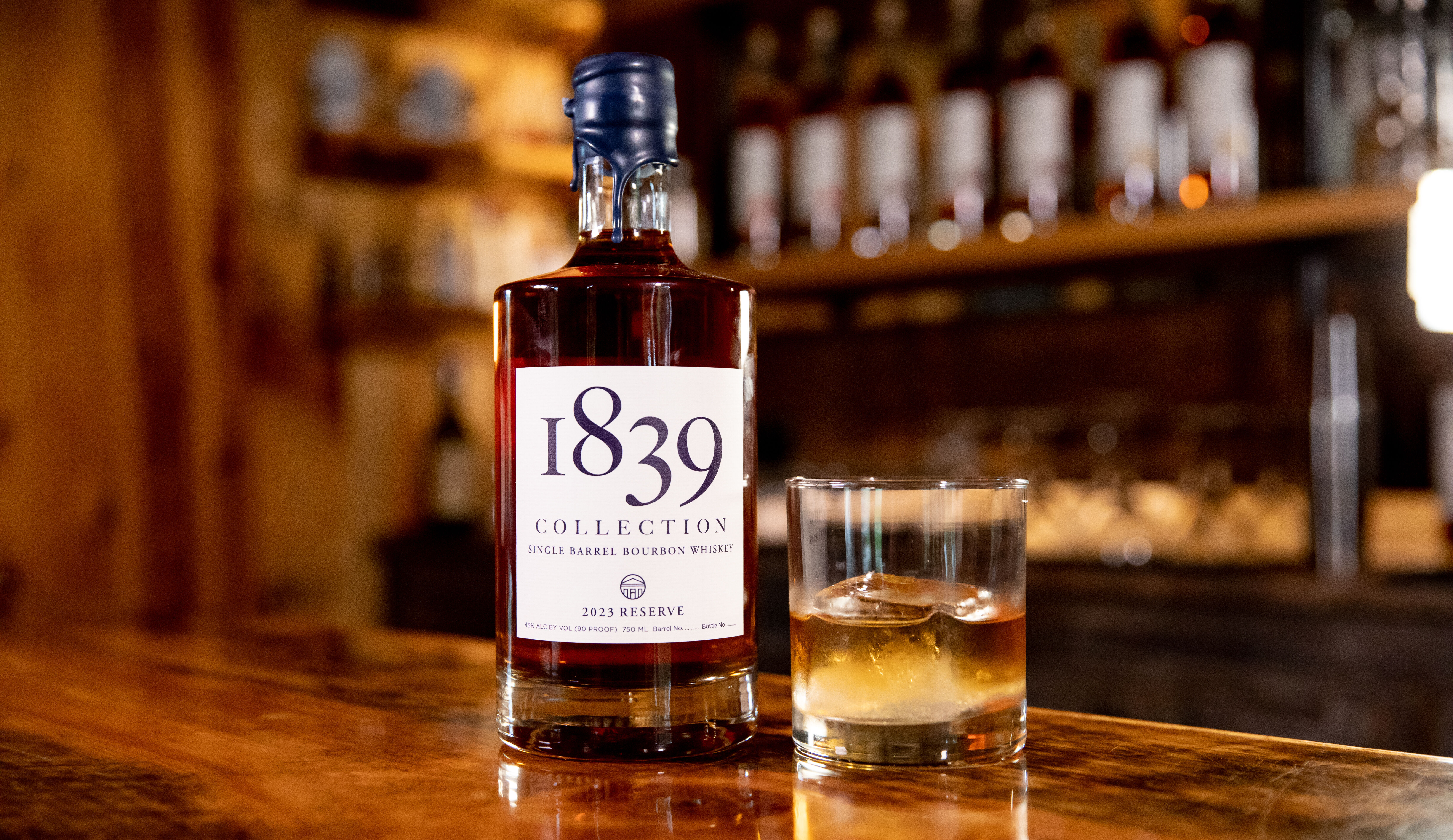 1839 Collection Bourbon Whiskey - A bottle of 1839 Collection single barrel bourbon stands on a wooden bar. The label reads “1839 Collection” and “2023 Reserve.” A glass of bourbon with a large ice cube sits beside the bottle. Shelves of additional bottles blur softly in the warm-lit background.
