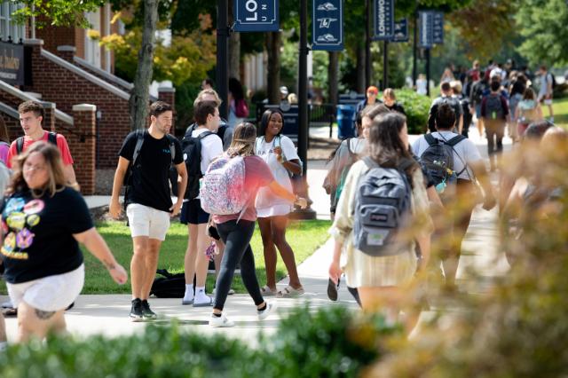 Students on Brock Commons - Students walk and talk along a tree-lined campus sidewalk. Many carry backpacks and wear casual clothing. Longwood banners hang from lampposts overhead. Brick buildings and green lawns frame the busy walkway.