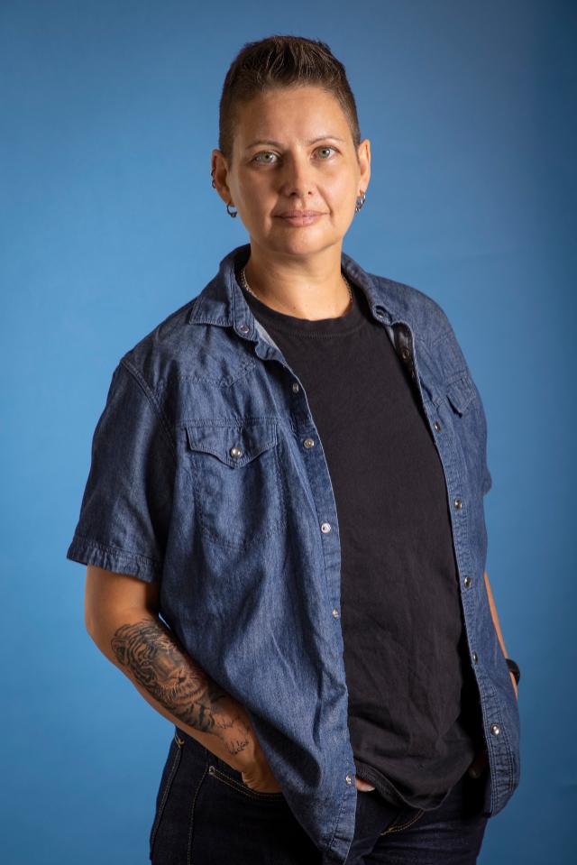 Carolina De Robertis, credit: Lori Eanes - Portrait of a person with short hair standing against a blue background. They wear a black shirt layered under a short-sleeve denim button-down. A tattoo is visible on their forearm, and their hands rest in their pockets. The subject faces the camera with a calm, confident expression.