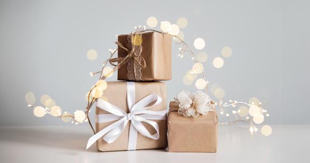 Pile of holiday gifts - Three brown paper–wrapped gift boxes stacked on a white surface. Boxes are tied with white ribbon, twine, and lace accents. Soft white string lights are draped around the presents. Warm bokeh lights glow in the background.