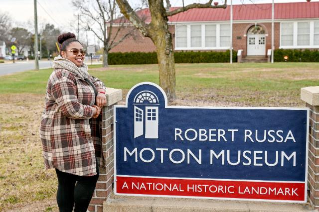 Jocelyn Watson ’24 - A woman stands beside a sign for the Robert Russa Moton Museum. The sign reads “A National Historic Landmark.” A brick building with a red roof sits in the background. She smiles outdoors on a grassy lawn near the street.