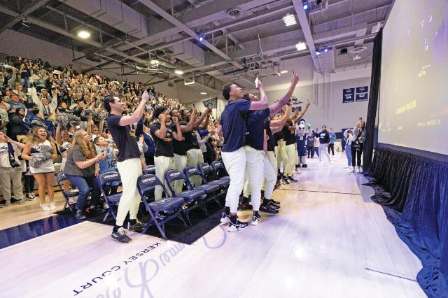 Selection Sunday - Longwood basketball players and fans react to an announcement on a large screen. Players stand from the bench cheering and holding up their phones. A packed crowd in the gym celebrates in the background. The scene captures an energetic moment during a team watch event.