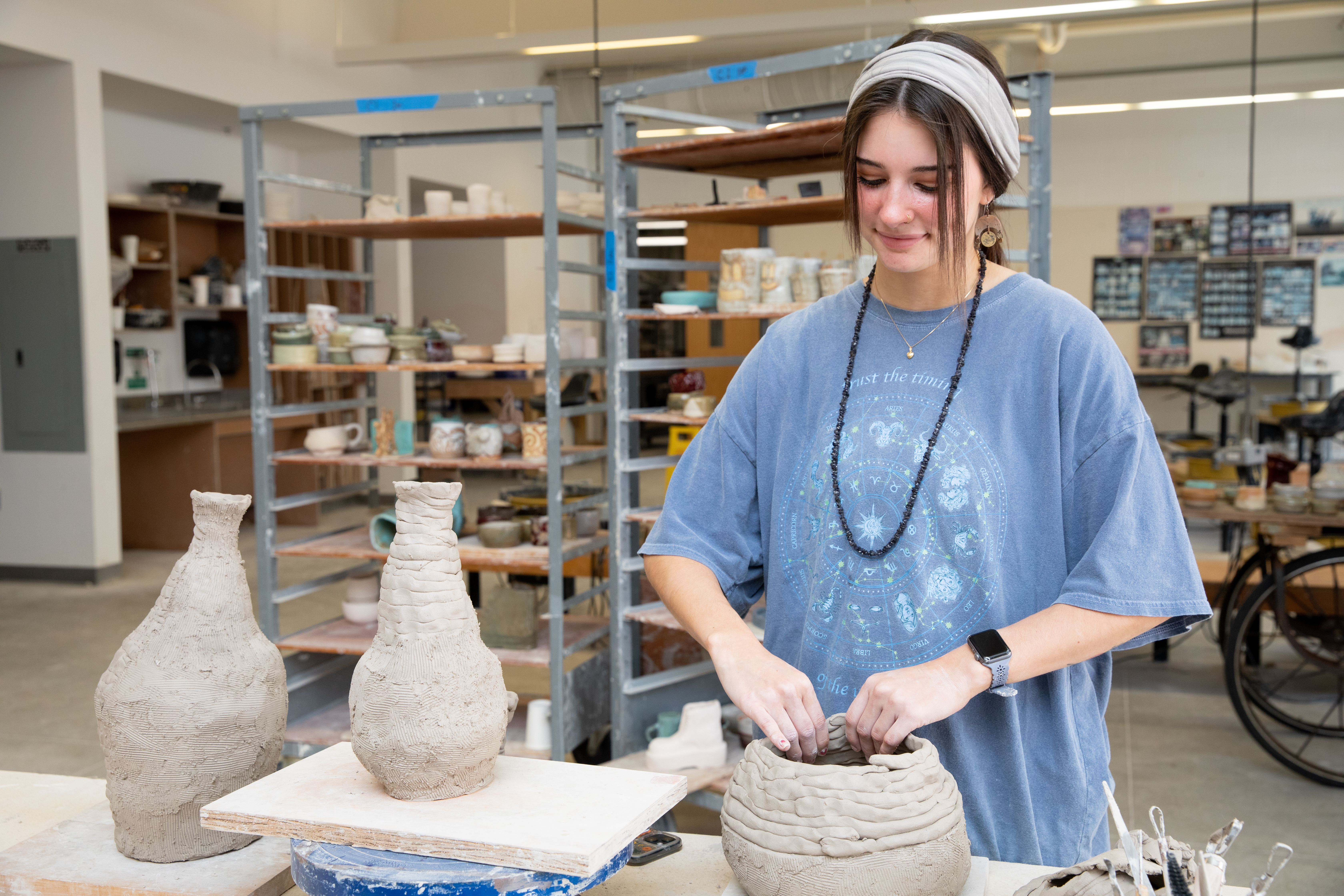 Student working in an art studio - A student shapes a clay vessel in a ceramics studio. Two textured clay vases sit on a nearby work surface. Shelving behind her holds finished pottery and supplies. The bright studio space features tables, tools, and natural light.