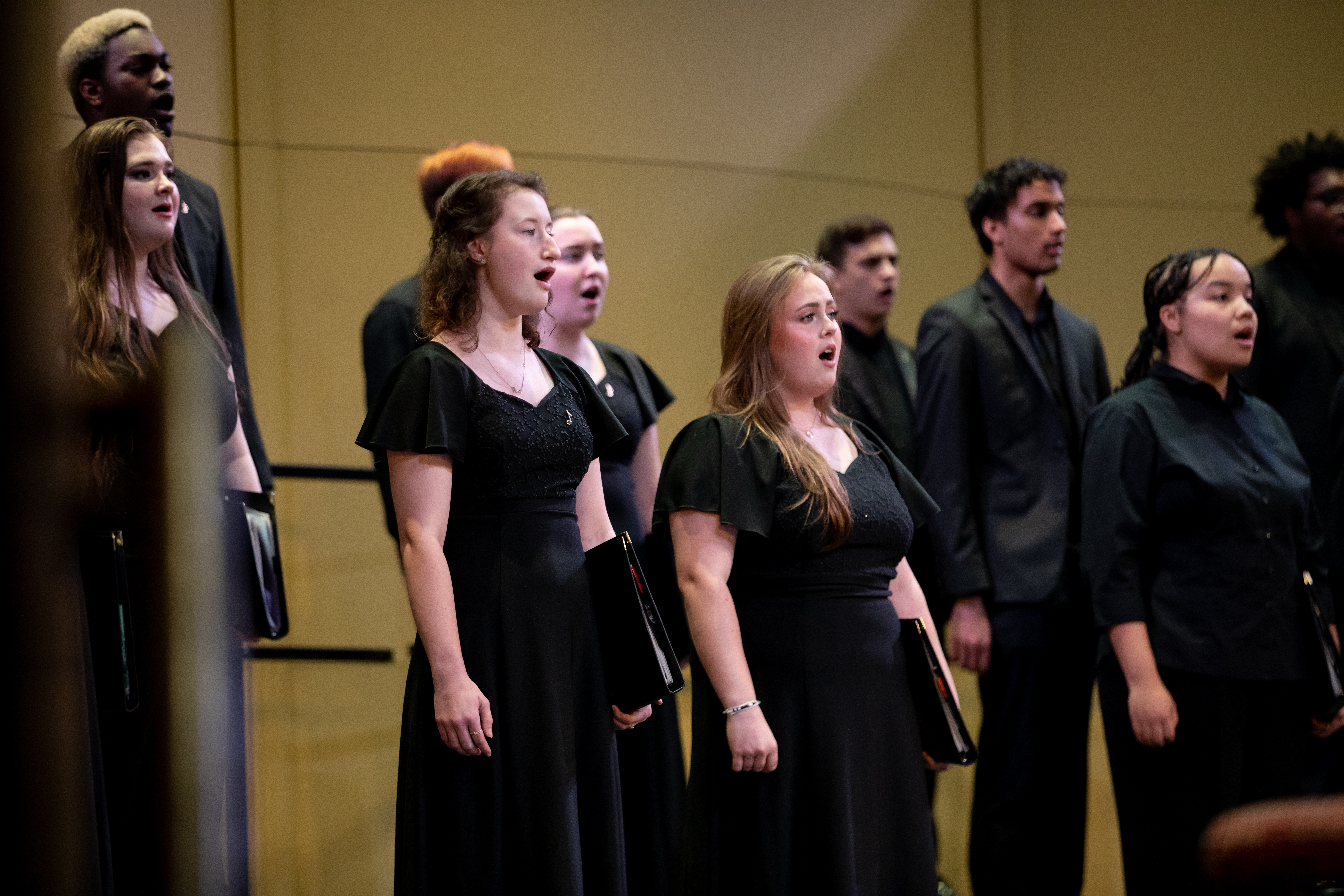 Choir in concert at Longwood - A group of students performs in a choral concert on stage. Singers dressed in black hold music folders and sing in harmony. Men and women stand in rows under warm stage lighting. The ensemble appears focused during a live performance.