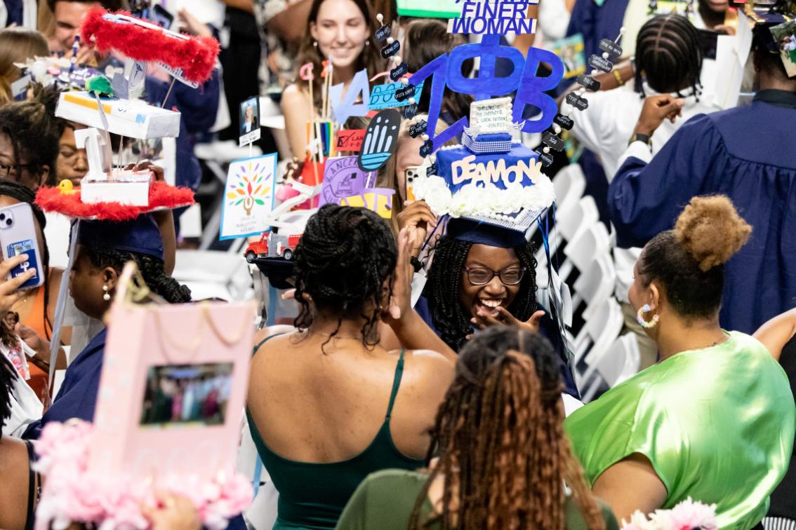 Students being capped at Convocation in the Joan Perry Brock Center - A graduate in cap and gown smiles while celebrating with friends. Her mortarboard is decorated with layered blue designs and the word “DEMON.” Other graduates wear colorful, creative cap decorations around her. The crowd gathers closely during a lively commencement ceremony.
