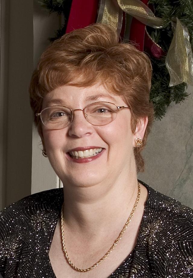 Diane Bottoms Boxley ’72 - Headshot of a smiling woman with short auburn hair and glasses. She wears gold earrings, a gold necklace, and a black patterned top. A holiday wreath with red ribbon is visible behind her. The portrait is warmly lit against an indoor background.